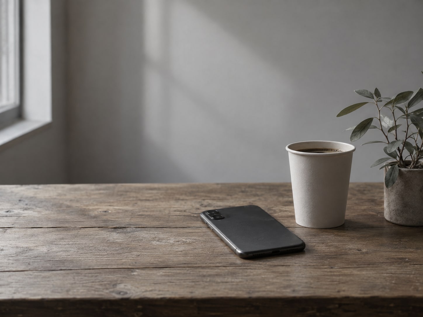 A phone lying face down beside a paper cup of coffee and a small plant on a wooden table.