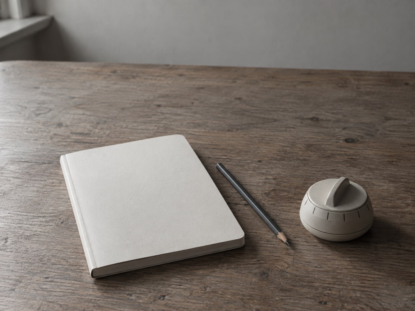 A closed notebook and a pencil beside a small kitchen timer on a wooden desk.