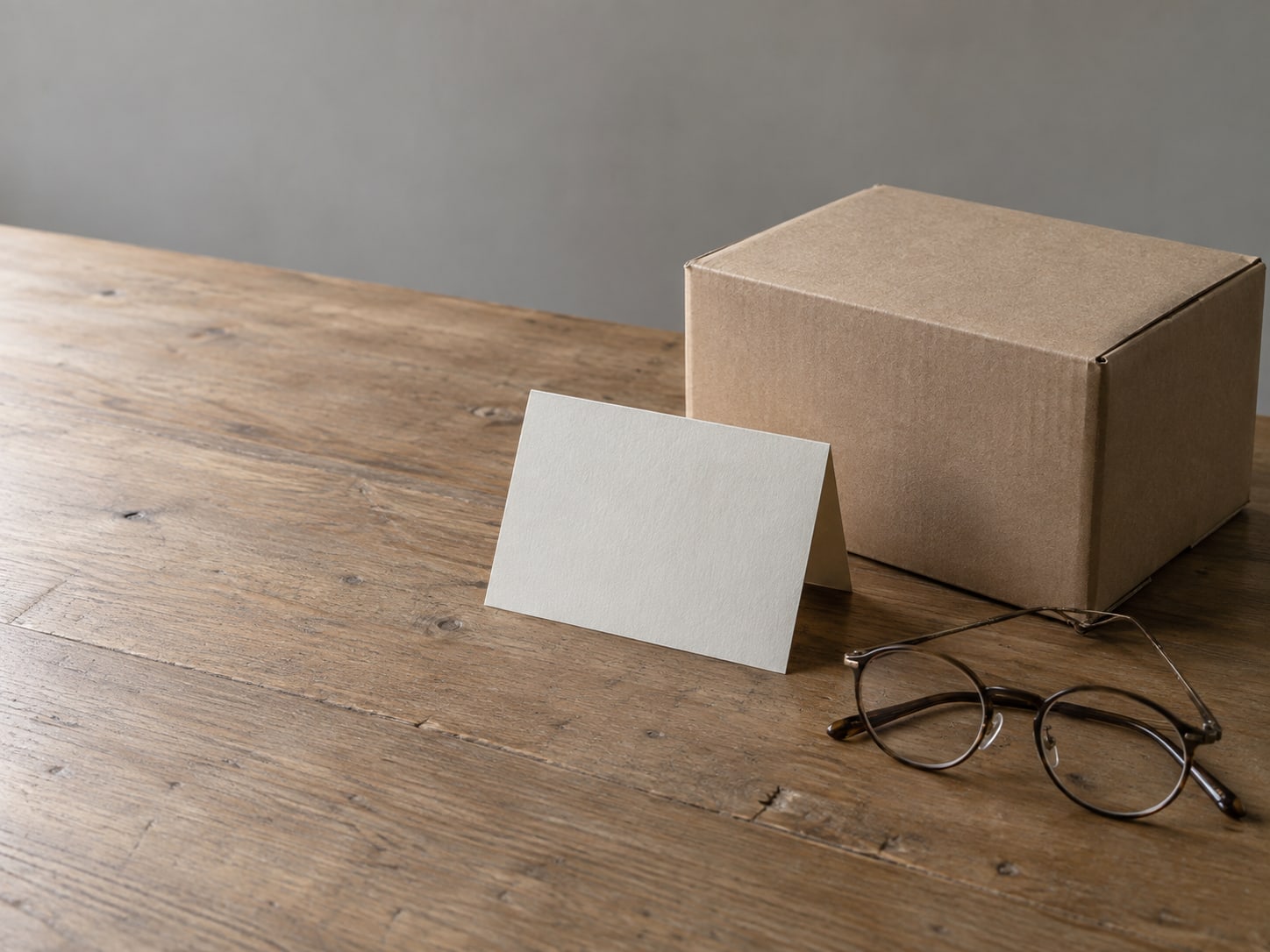 A sealed cardboard box beside a blank folded card and a pair of reading glasses on a desk.