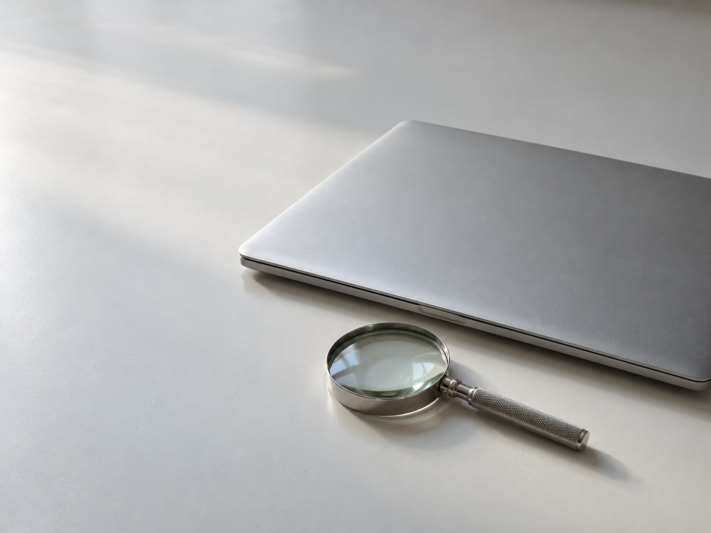 Una laptop cerrada junto a una pequeña lupa sobre un escritorio blanco liso.