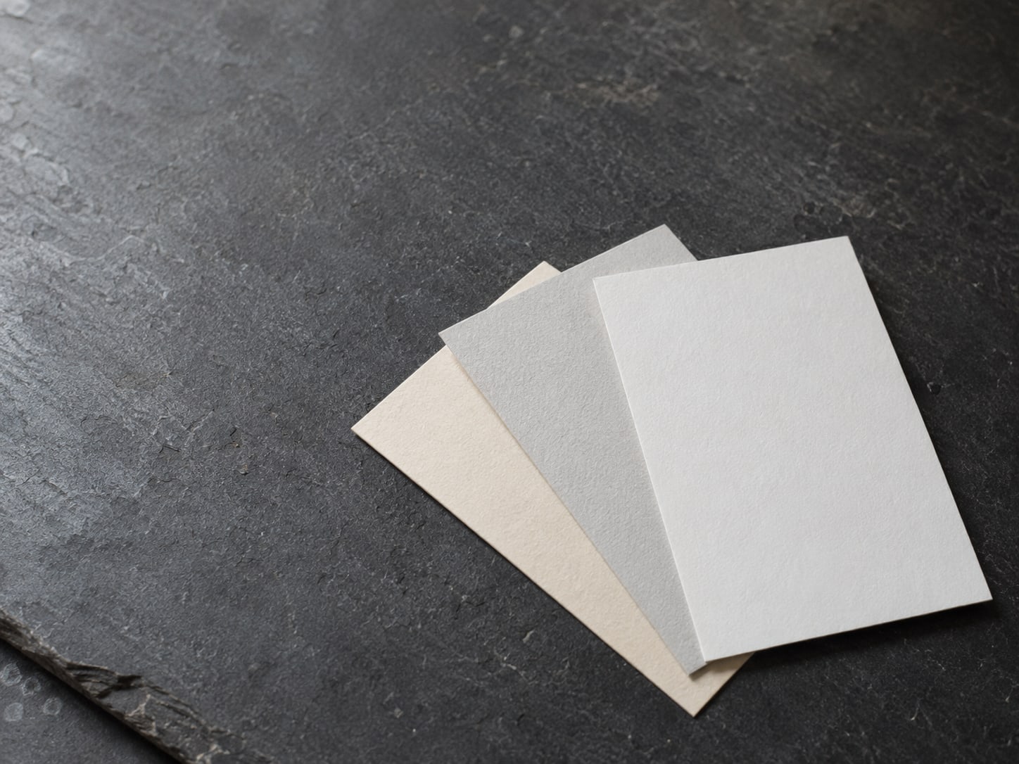 Tres tarjetas de papel en blanco desplegadas sobre una superficie de pizarra, cada una de un tono ligeramente distinto.