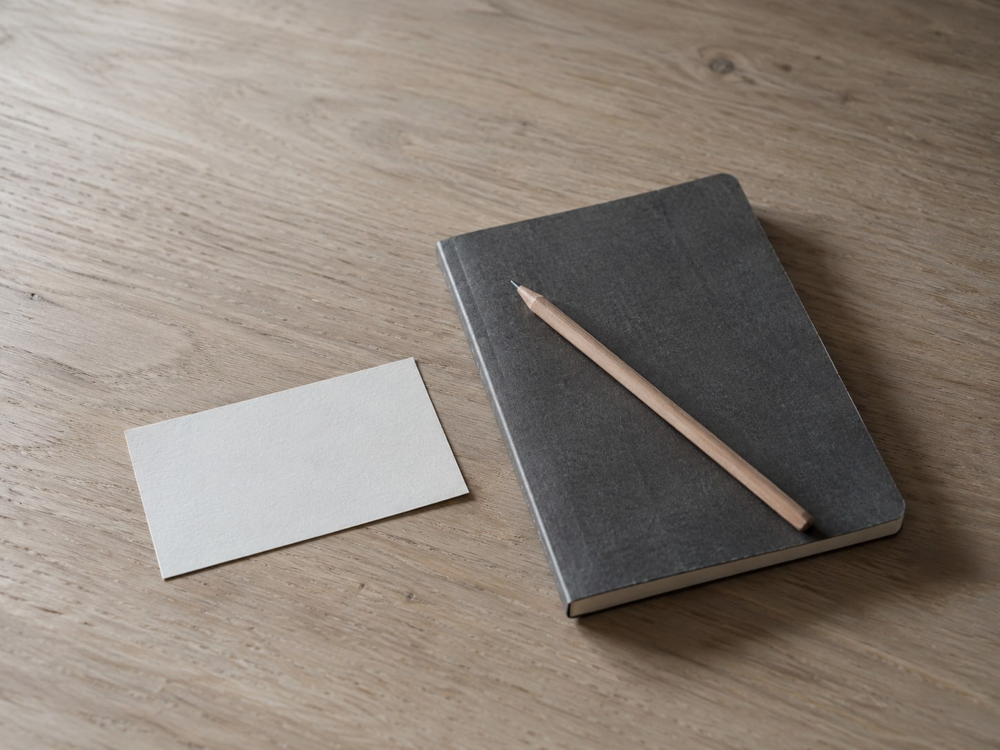 A closed notebook with a pencil resting across it beside a blank index card on a wooden desk.