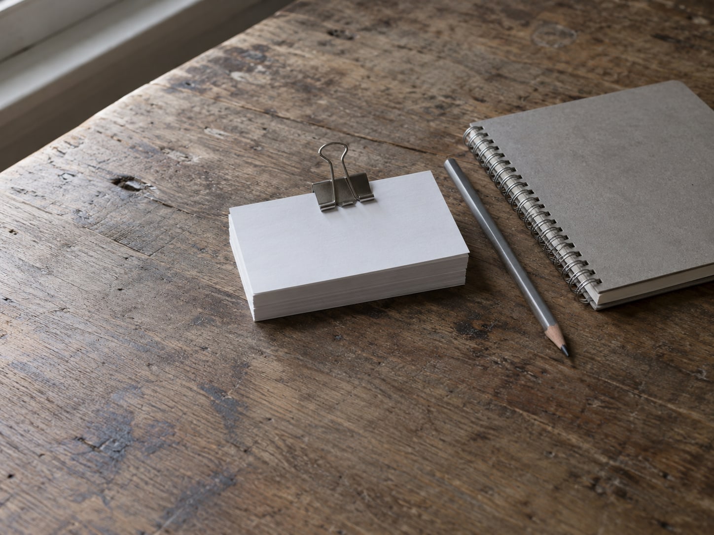A stack of blank index cards held by a binder clip beside a pencil and a closed spiral notebook.