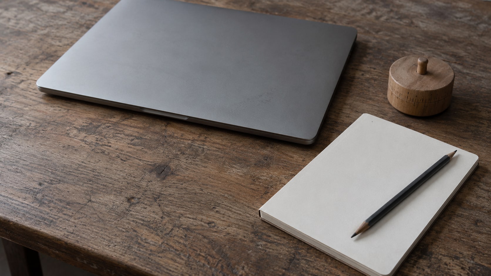 A closed laptop, a wooden kitchen timer and a blank notepad with a pencil on a worn desk.