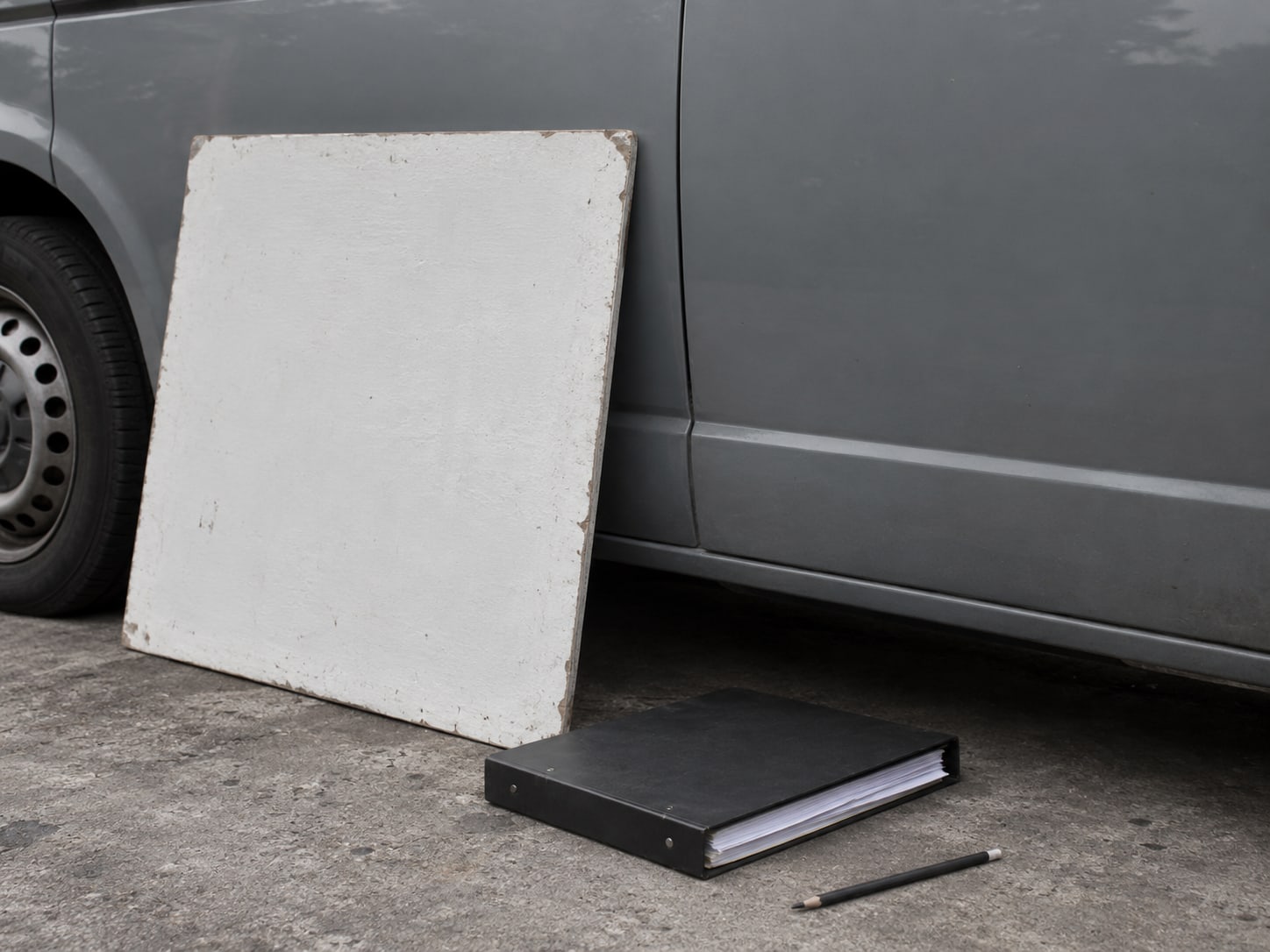 A plain painted work sign leaning against a van door beside a closed binder of blank invoices and a pencil.