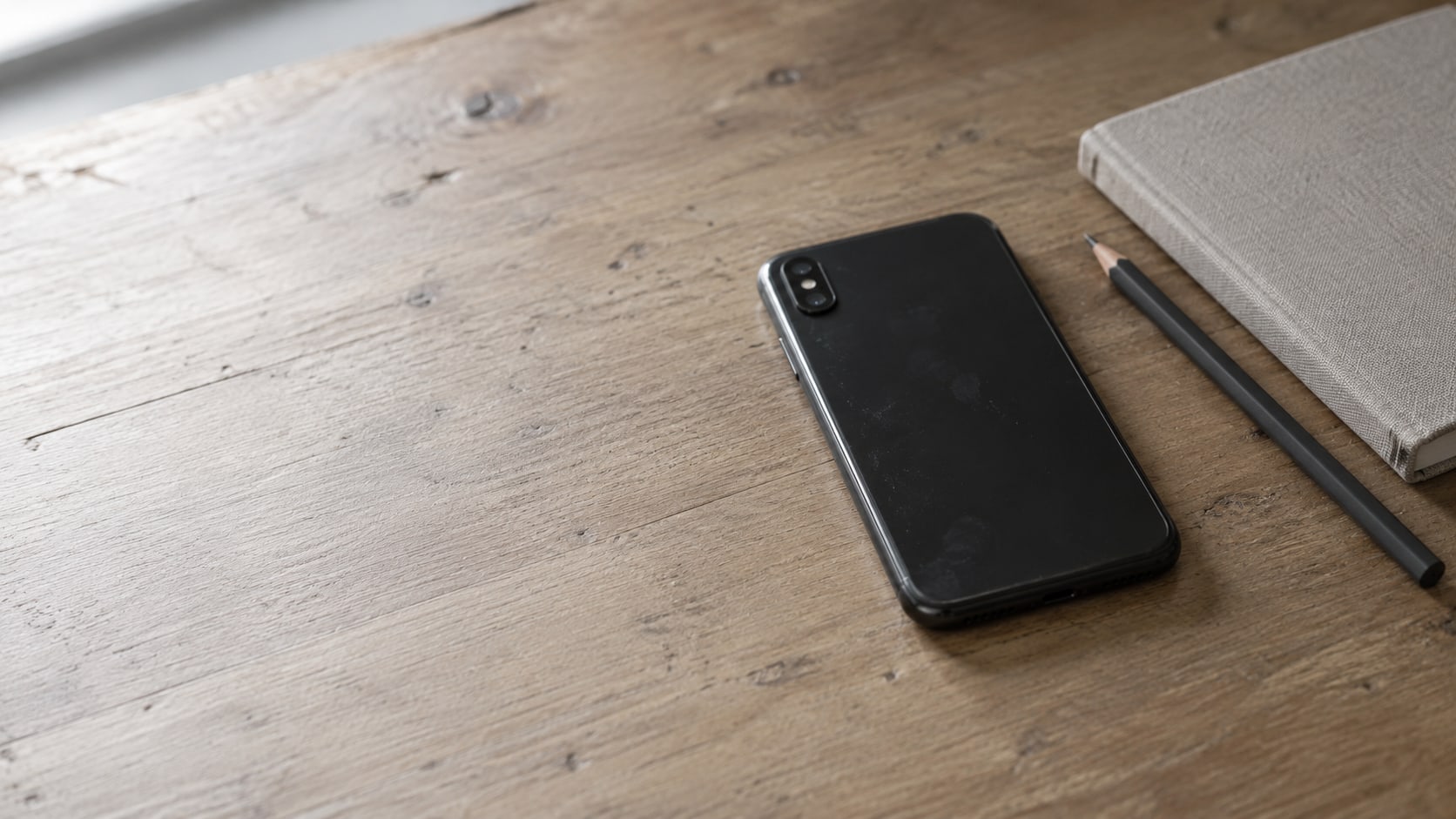 A smartphone lying face down on a wooden desk beside a closed notebook and a pencil.