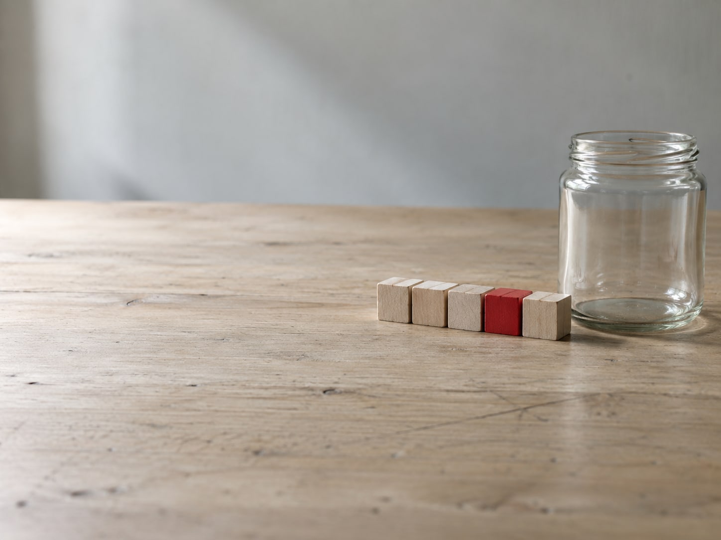 A neat row of small wooden blocks, one painted bright red, beside an empty glass jar on a pale desk.