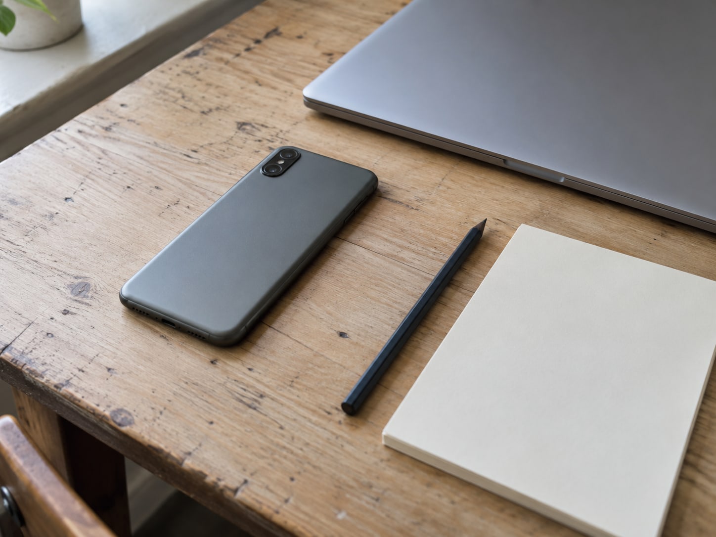 A closed laptop and a smartphone lying face down beside a pencil and a blank notepad on a wooden desk.
