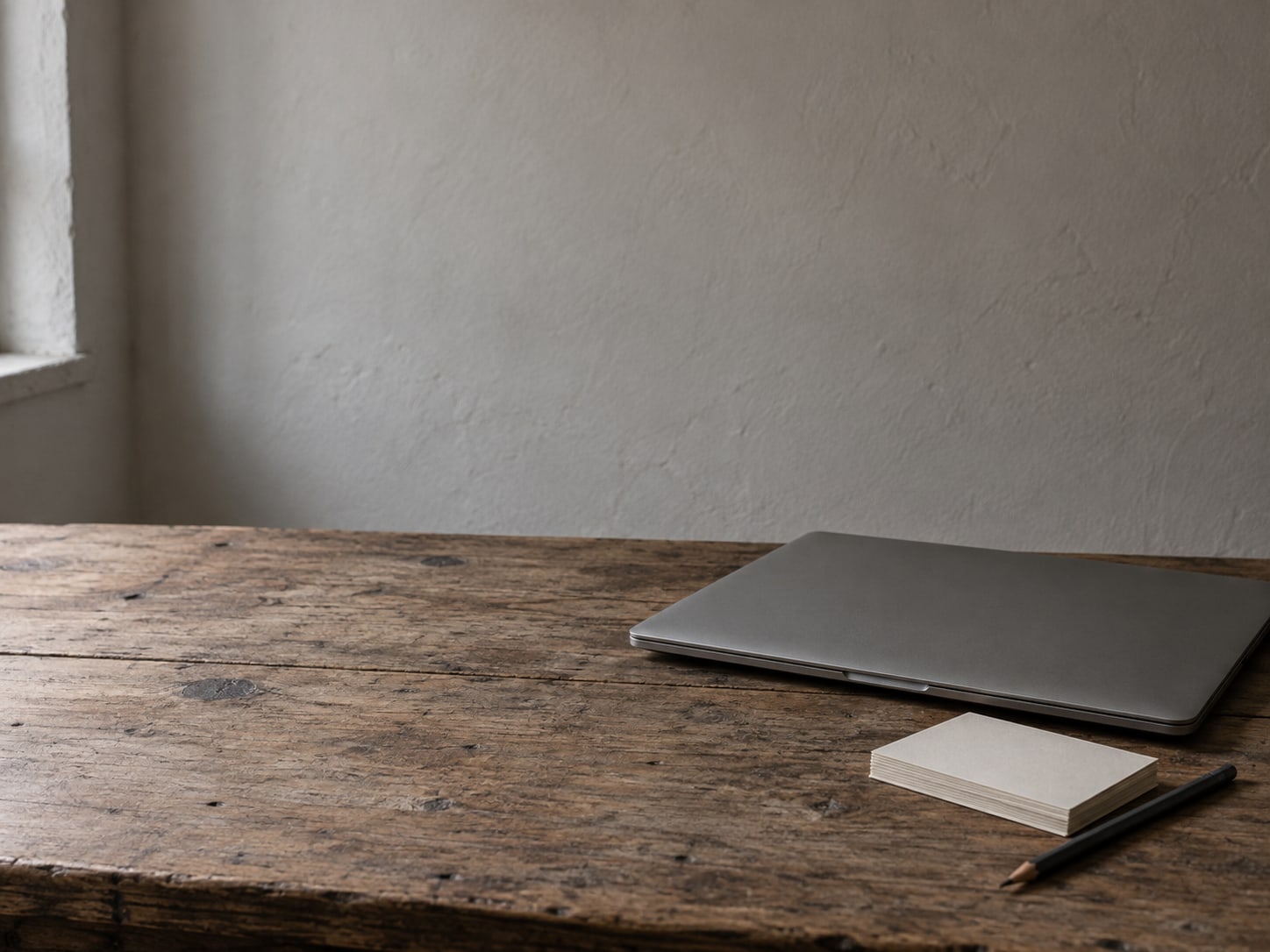 A closed laptop beside a small stack of blank index cards and a pencil on a worn wooden desk.