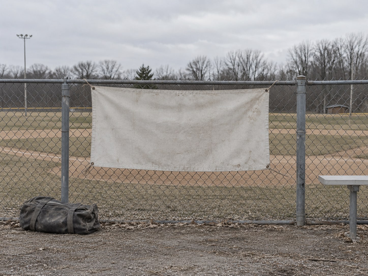 A blank canvas banner tied to a chain-link fence beside a canvas equipment bag on a baseball field.