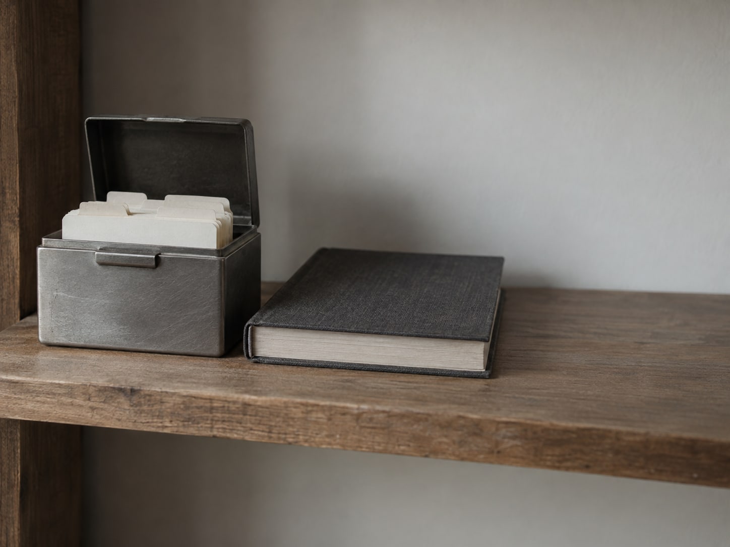 A small metal file box with blank tabbed dividers on a wooden shelf, lid open, next to a closed ledger.
