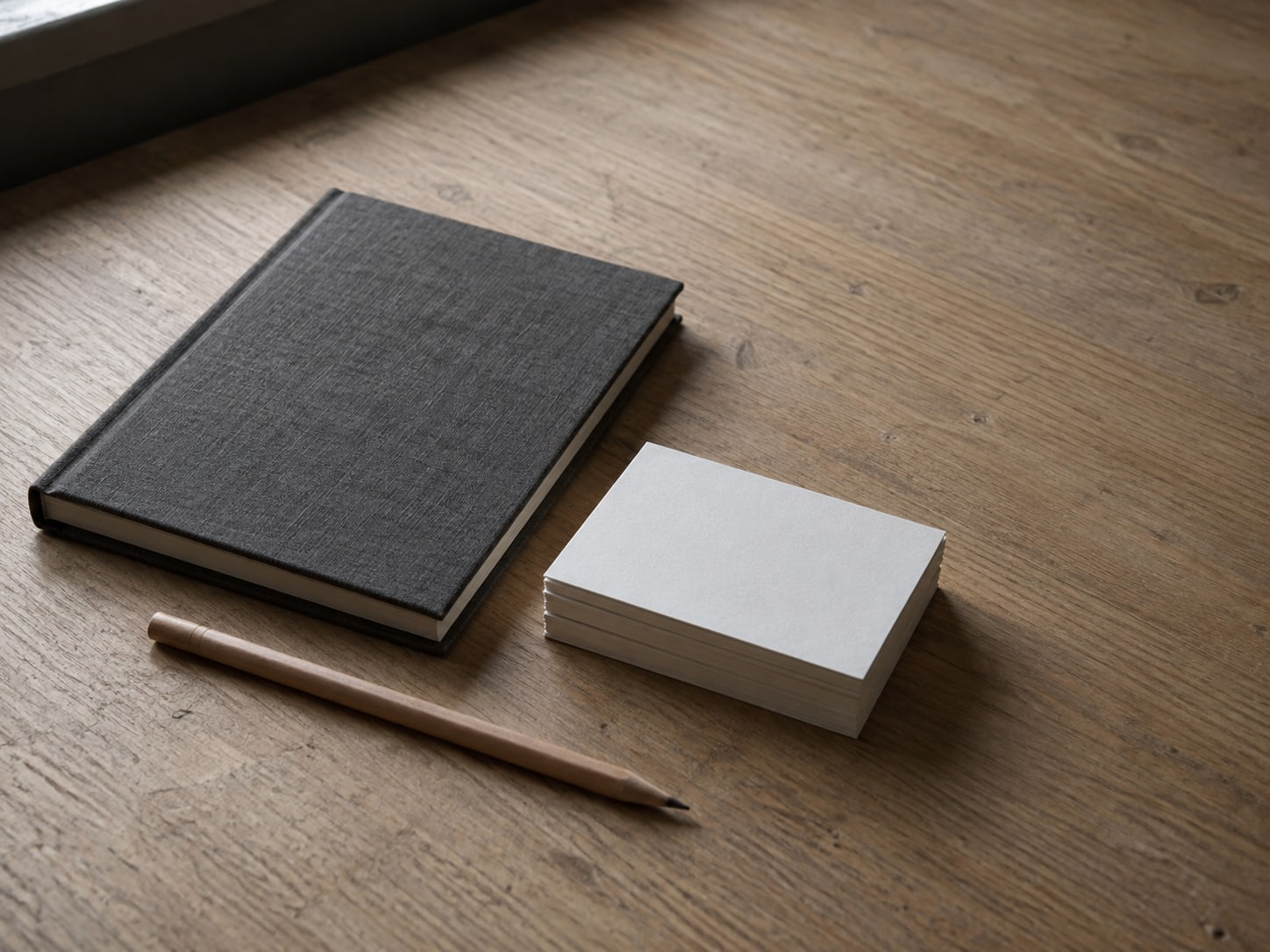 A small stack of blank index cards squared neatly beside a closed notebook and a pencil on a light oak desk.
