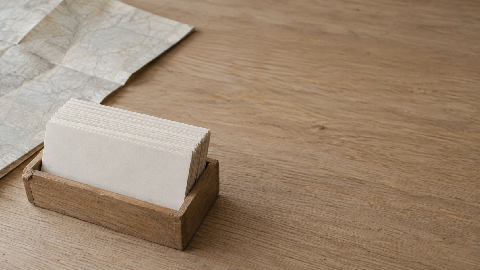 A neat row of blank index cards in a small wooden card tray beside a folded paper map on a light oak desk.