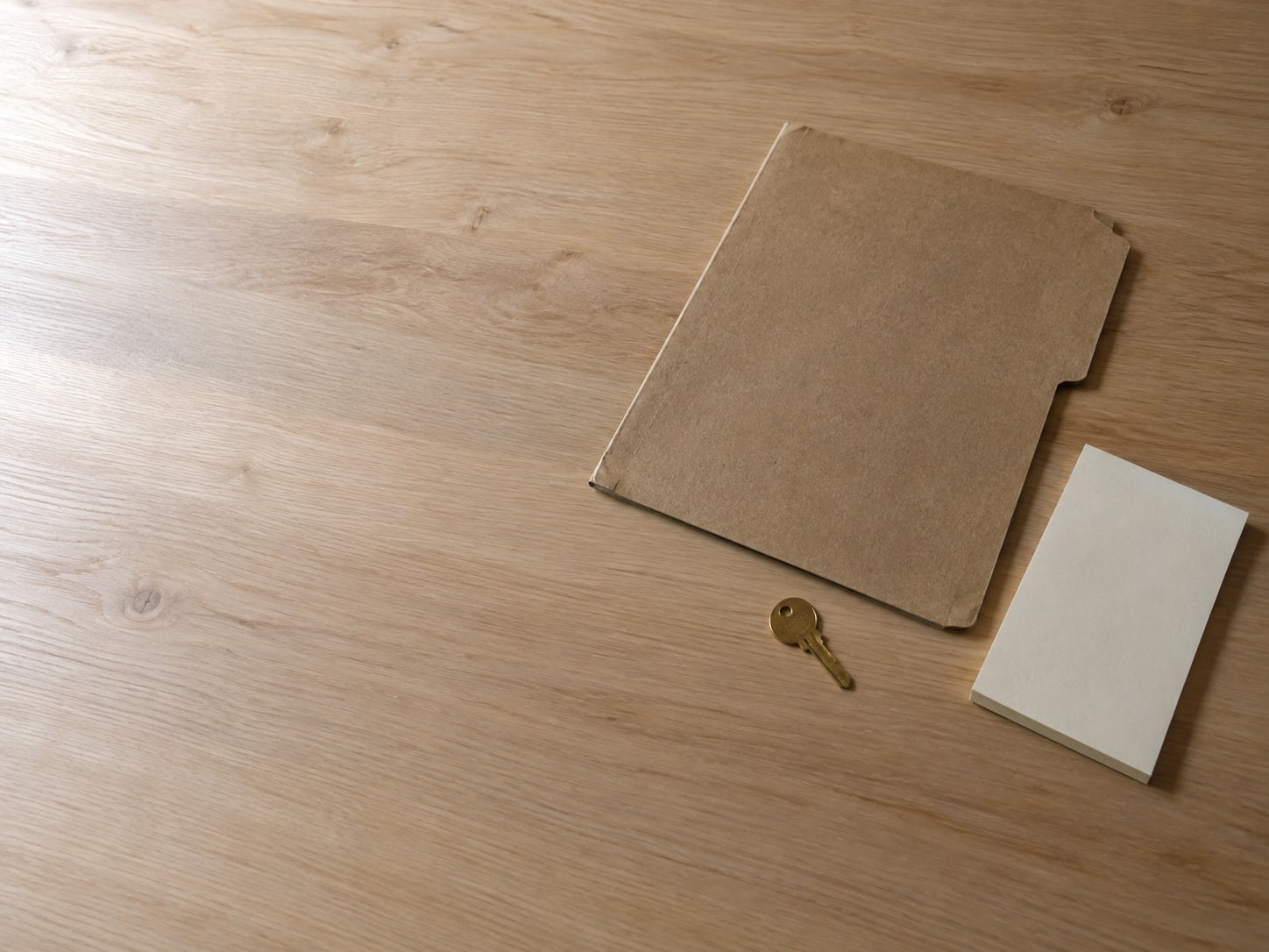 A closed brown folder and a small brass key lying on a clean wooden desk beside a blank notepad.