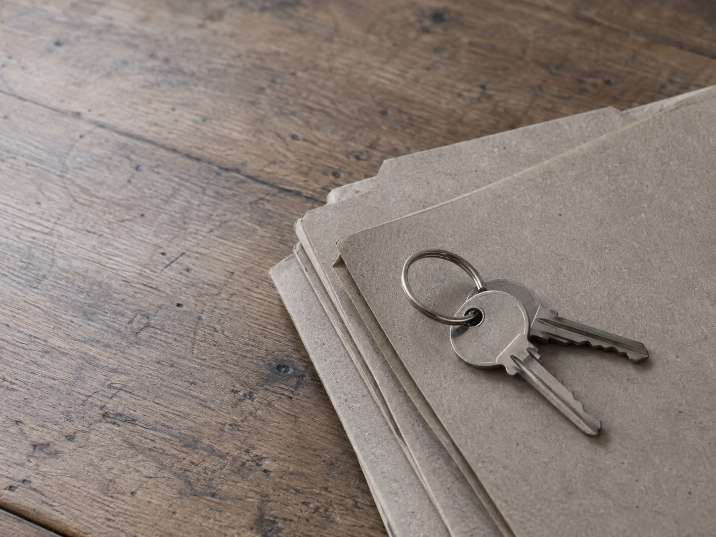 Two plain metal keys on a ring resting on a stack of unmarked cardboard folders on a desk.