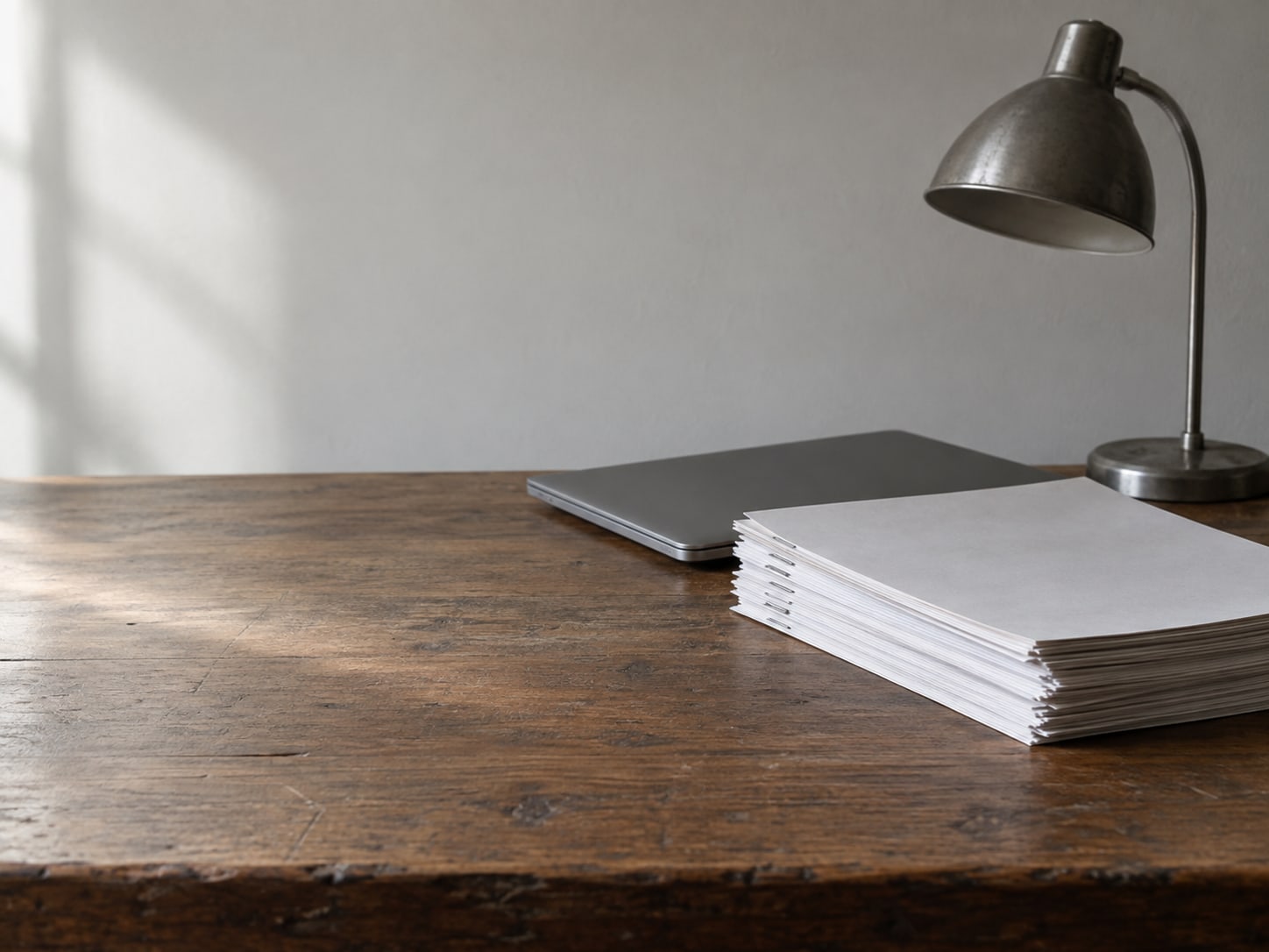 A neat pile of blank stapled papers next to a closed laptop and a small desk lamp on a wooden table.