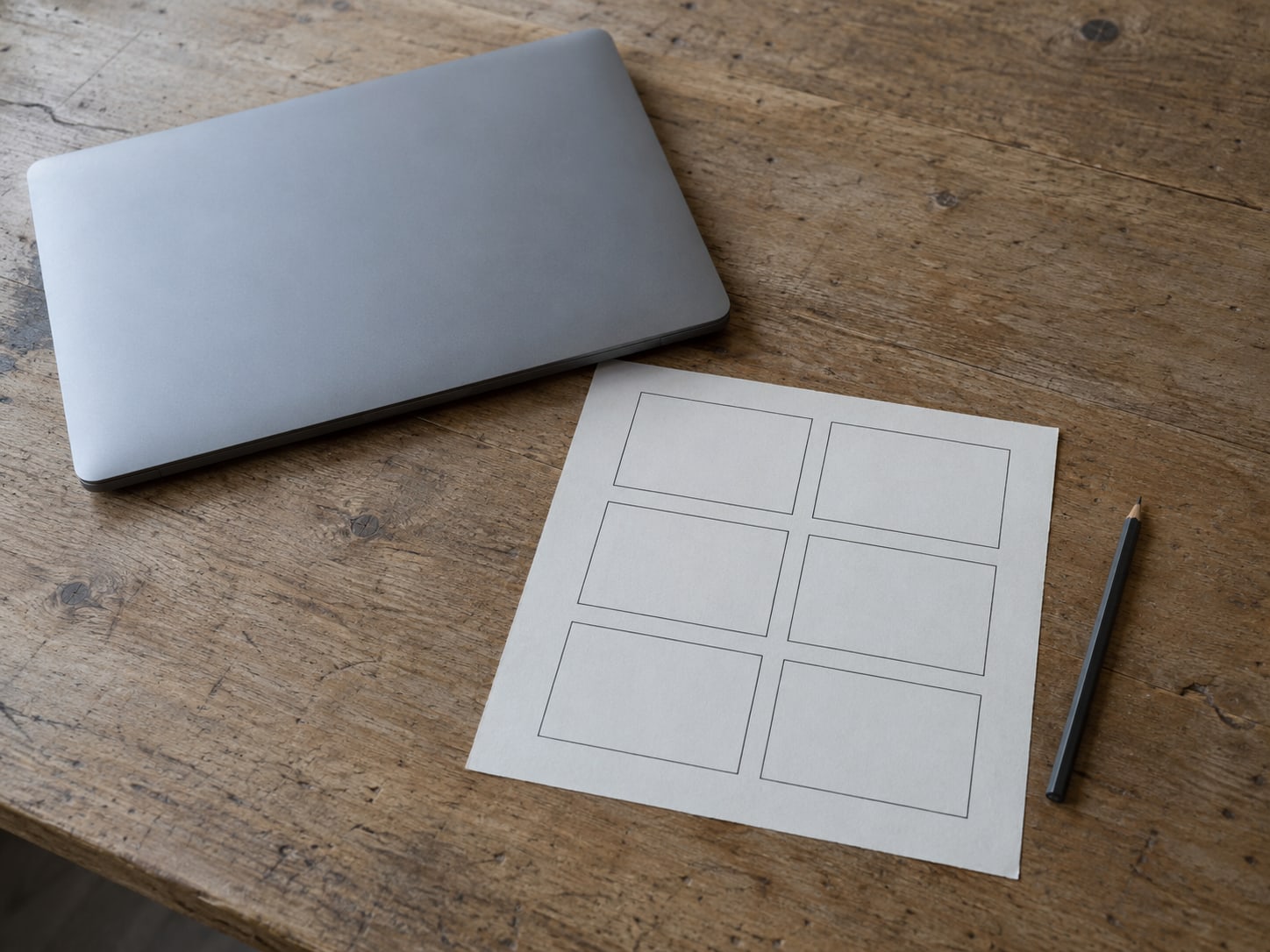 A closed laptop beside a blank paper storyboard of six empty frames and a pencil on a wooden desk.