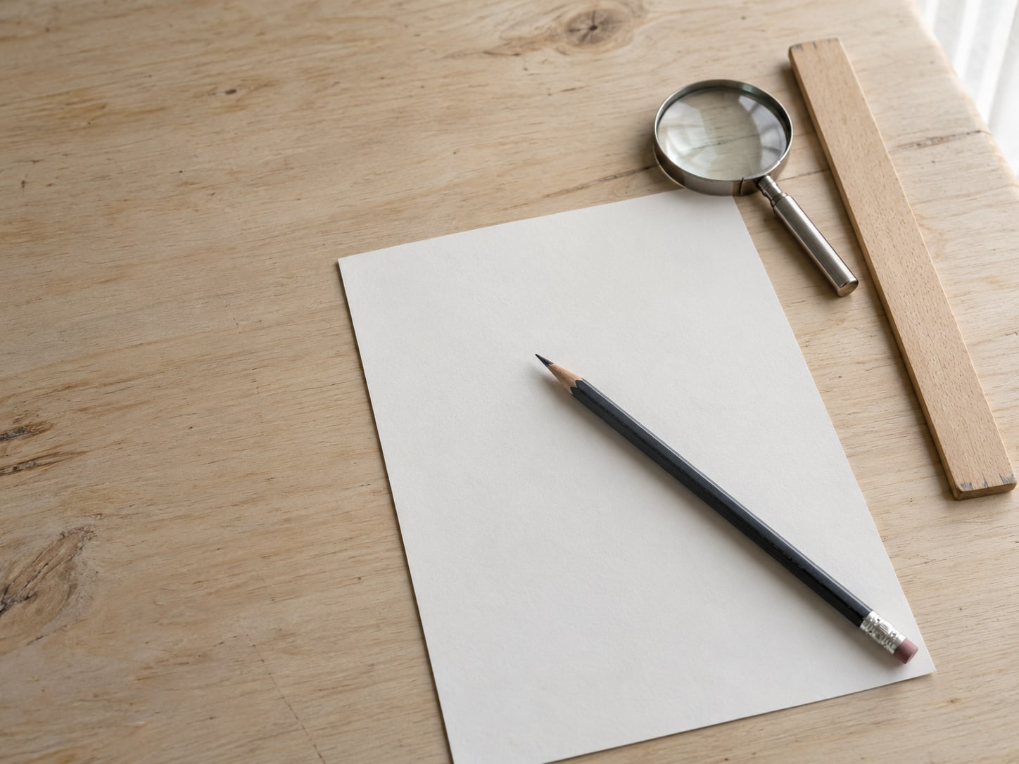 A pencil lying across a blank checklist sheet next to a small magnifying glass and a ruler on a wooden table.