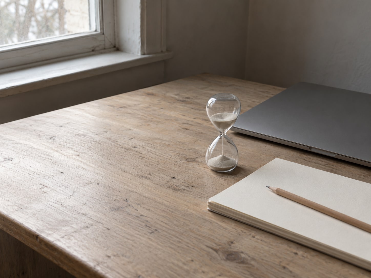 A closed laptop beside a small sand timer and a pencil resting on a blank notepad on a wooden desk.