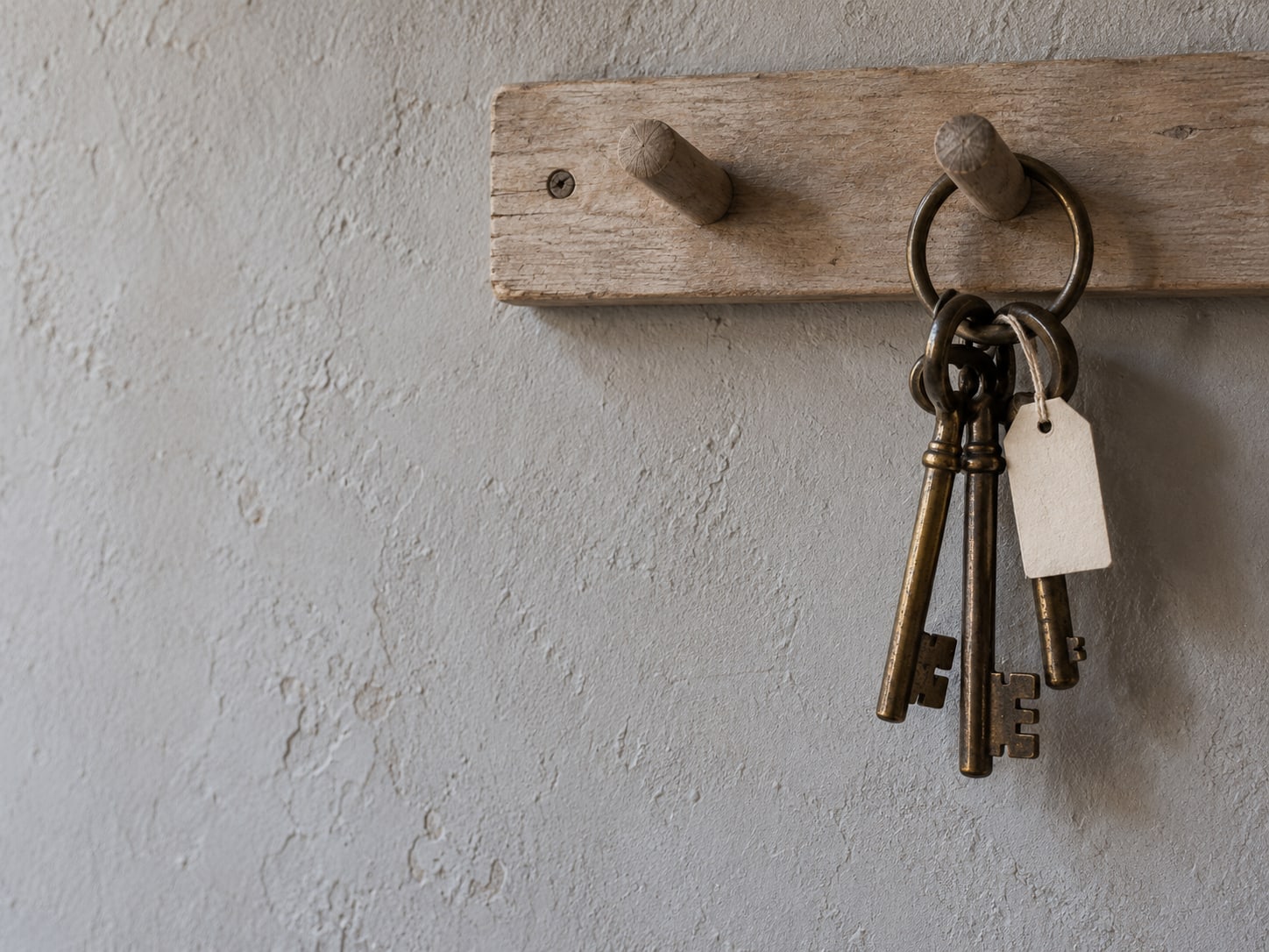 A ring of unmarked brass keys hanging from a wooden hook board, with a single key wearing a small blank tag.