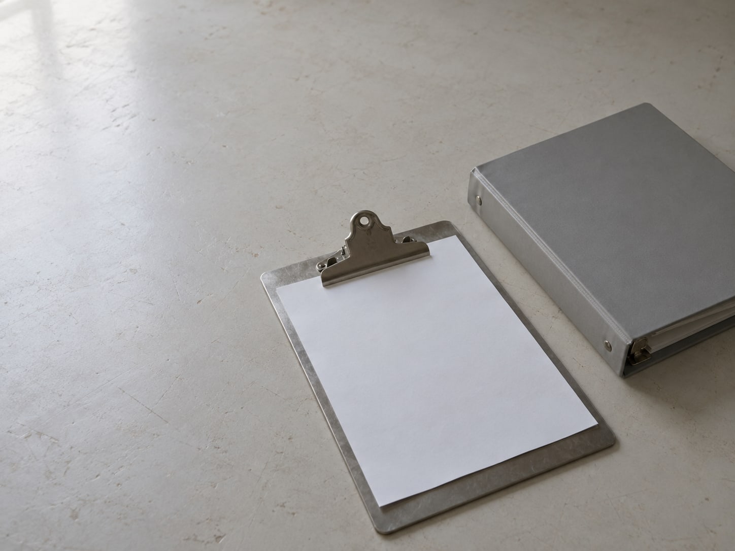 A clipboard with a blank sheet of paper beside a closed three-ring binder on a plain table.
