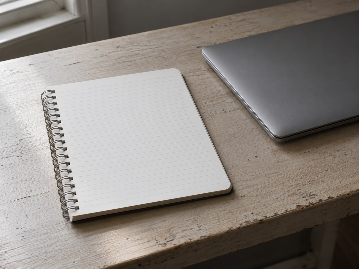 Una libreta de espiral abierta, con una columna de filas en blanco escritas a mano, junto a una laptop cerrada sobre un escritorio de madera liso.