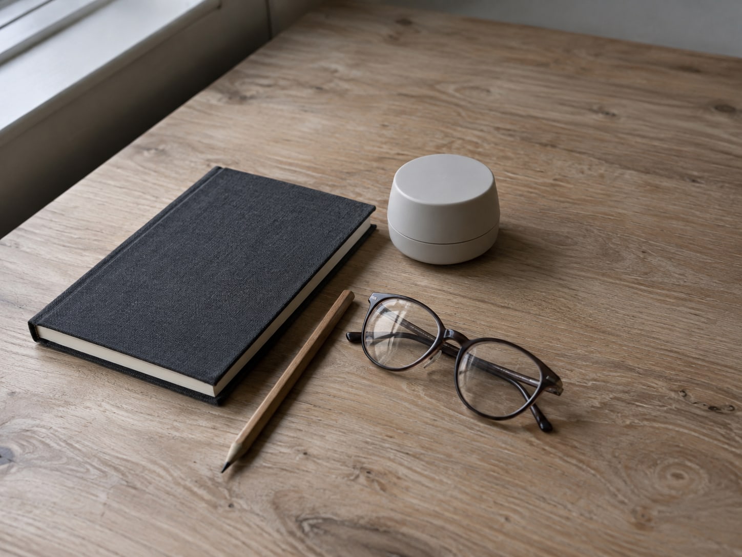 A small wooden desk with a closed notebook, a pencil and a plain round kitchen timer beside a folded pair of reading glasses.