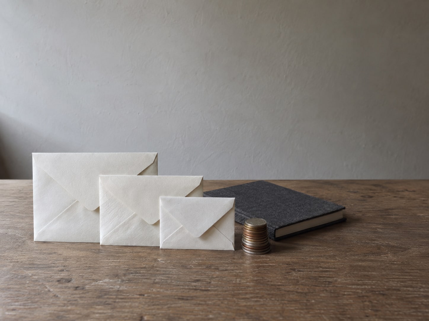 Three plain paper envelopes of different sizes lined up on a desk beside a small stack of coins and a closed notebook.