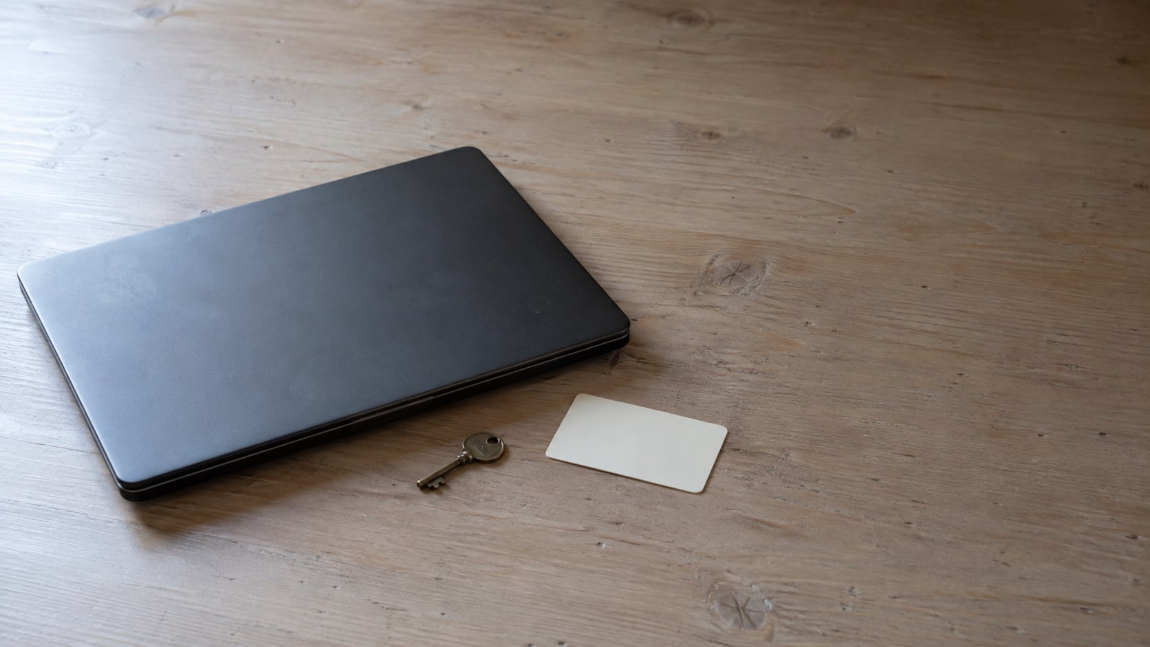 A closed laptop beside a small brass key and a blank membership card on a light wooden desk.