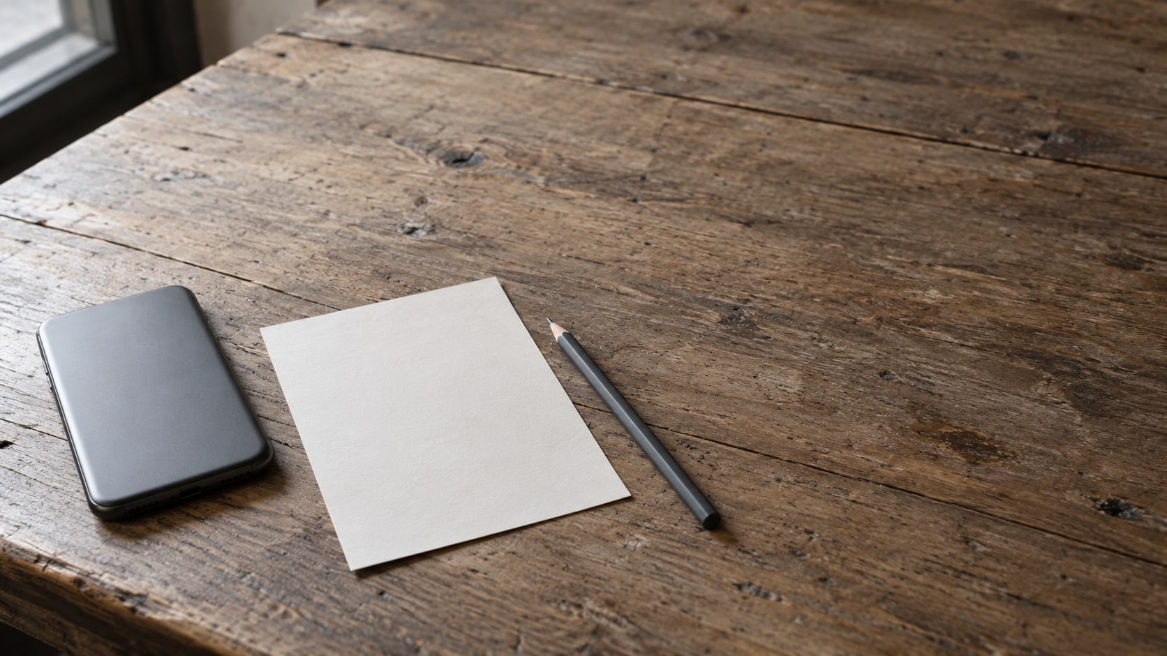A smartphone lying face down on a wooden counter beside a blank printed menu card and a pencil.