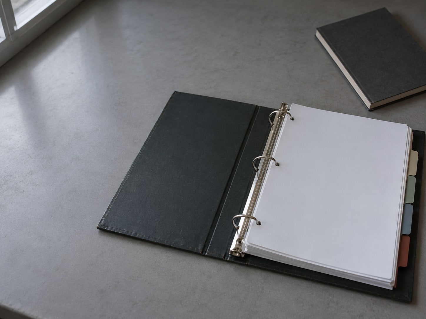 A three-ring binder of blank printed sheets with colored tab dividers on a desk beside a closed notebook.