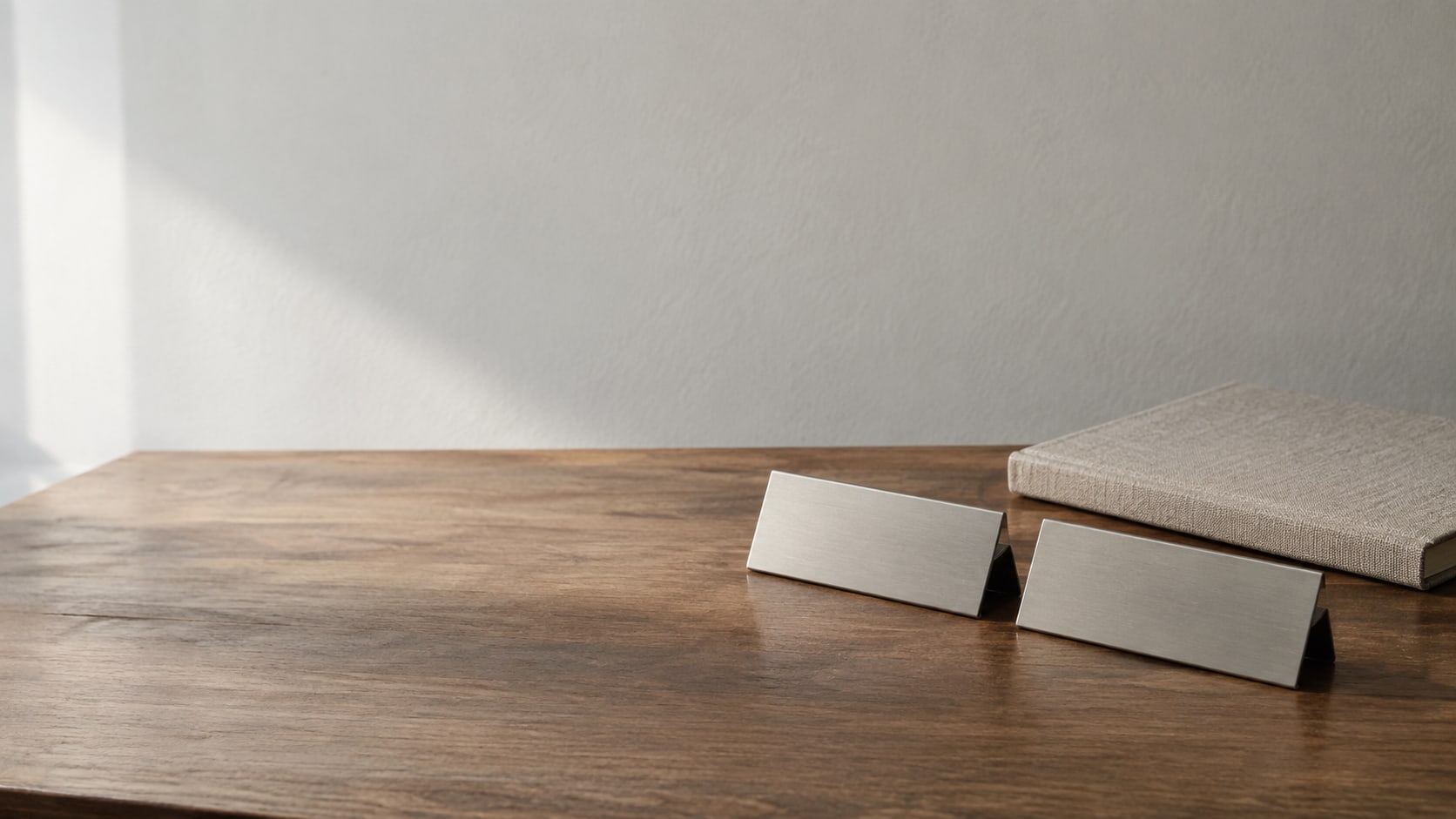 Two matching blank nameplates side by side on a wooden desk beside a closed notebook.