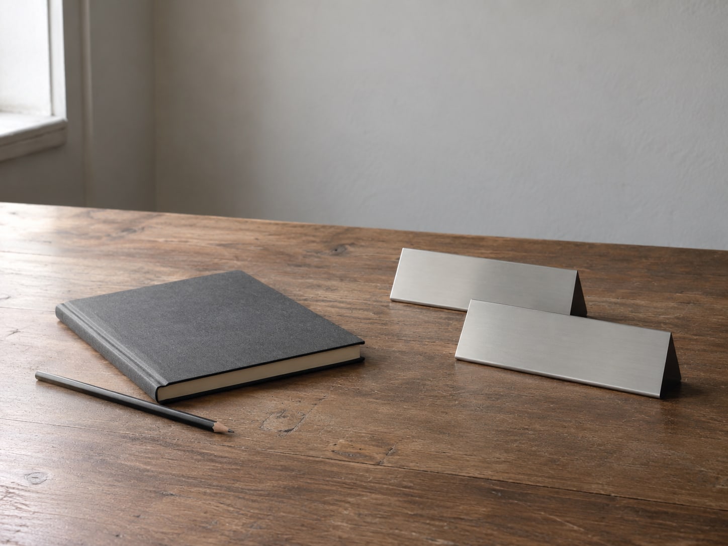 A closed notebook and a pencil beside two identical blank nameplates on a wooden desk.