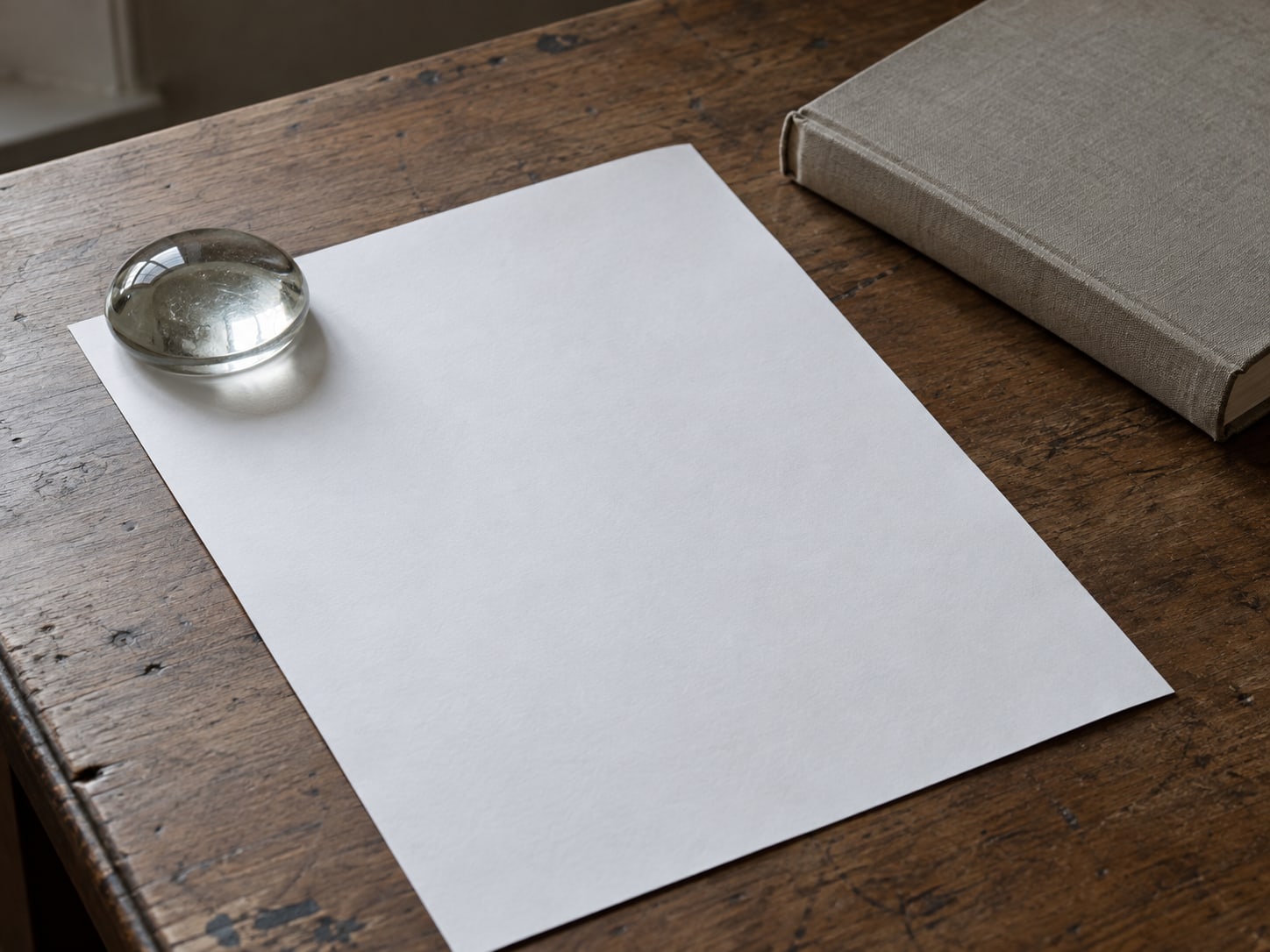 A single sheet of white paper pinned flat under a glass paperweight on a desk with a closed hardcover book nearby.