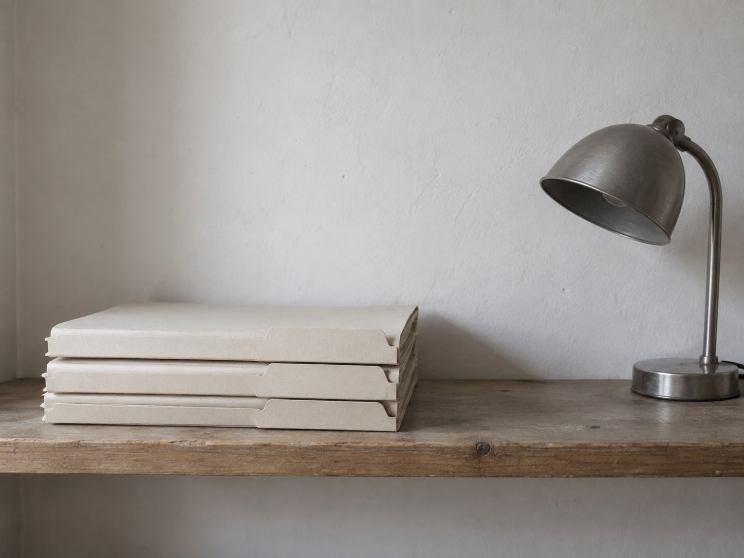A tidy stack of three folders with blank tabs sits on a wooden shelf beside a small desk lamp that is switched off.