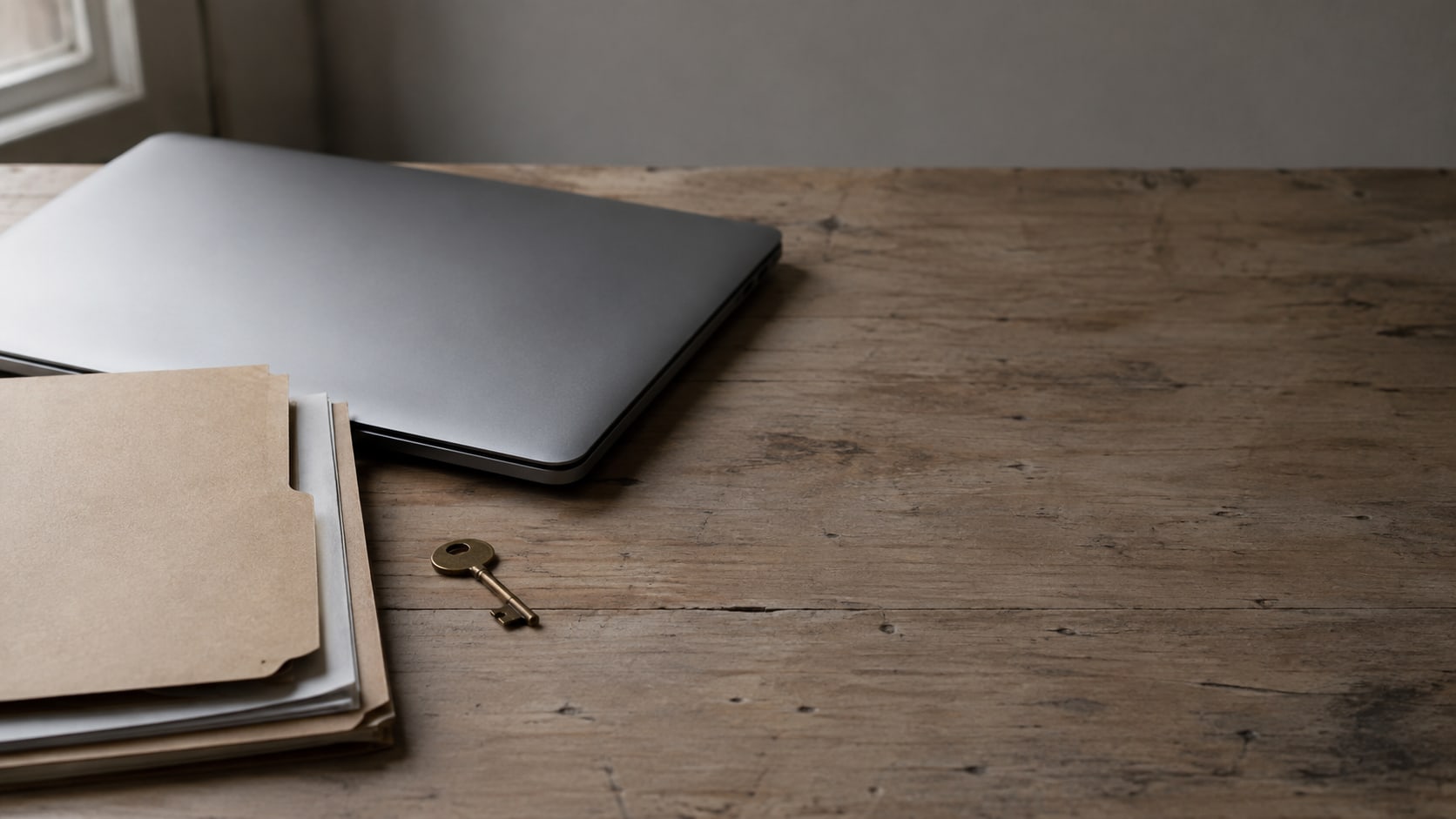 A closed laptop, a small brass key and a stack of blank folders on a plain wooden desk.