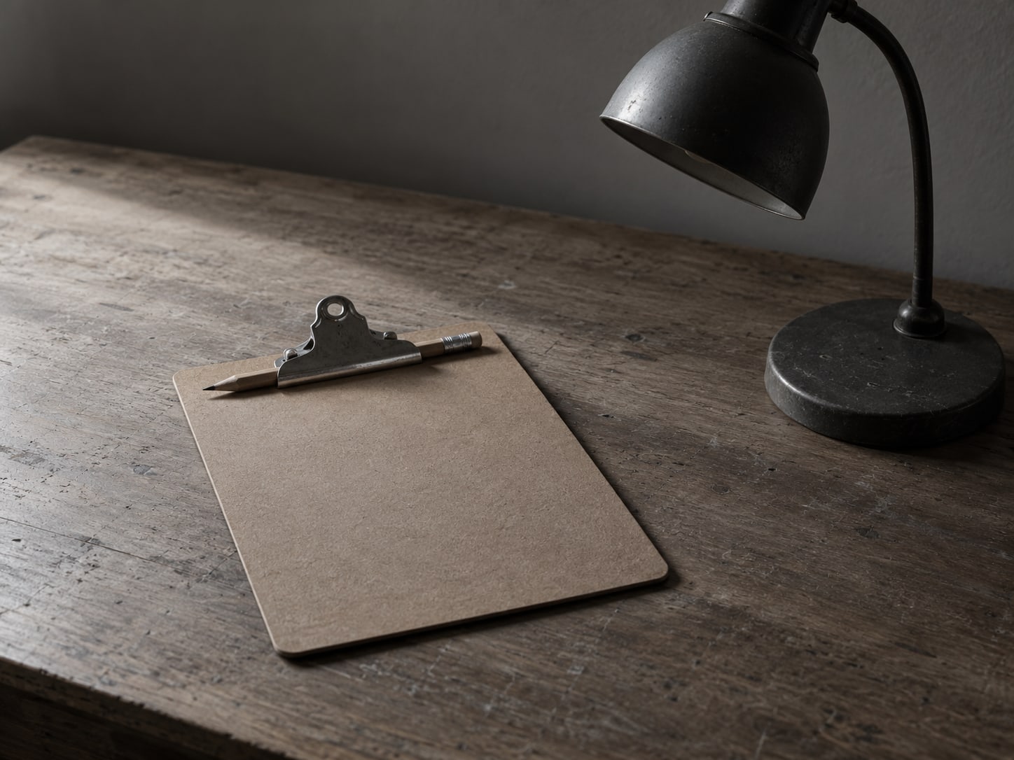 A closed clipboard with a pencil clipped to it, lying beside a small unlit desk lamp.