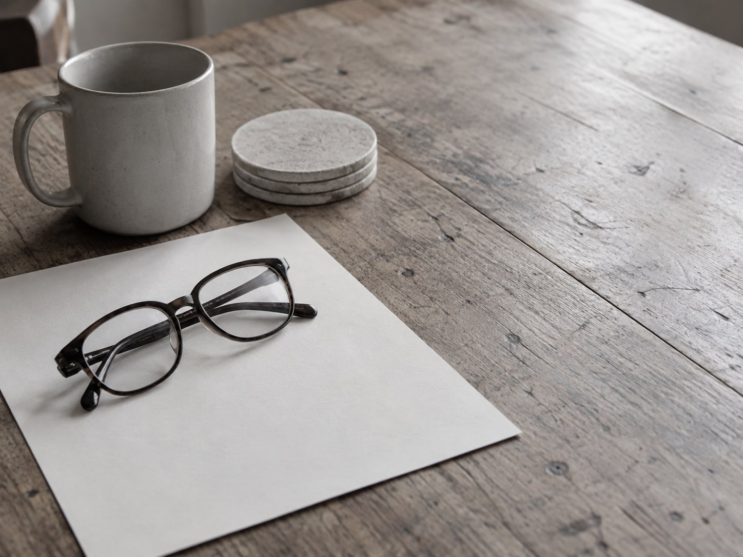 A pair of reading glasses folded on a printed page of blank lined paper next to a mug and a small stack of coasters.