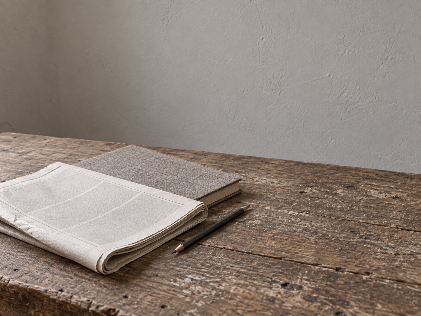 Un periódico doblado con columnas en blanco junto a un cuaderno cerrado y un lápiz sobre una mesa de cocina.
