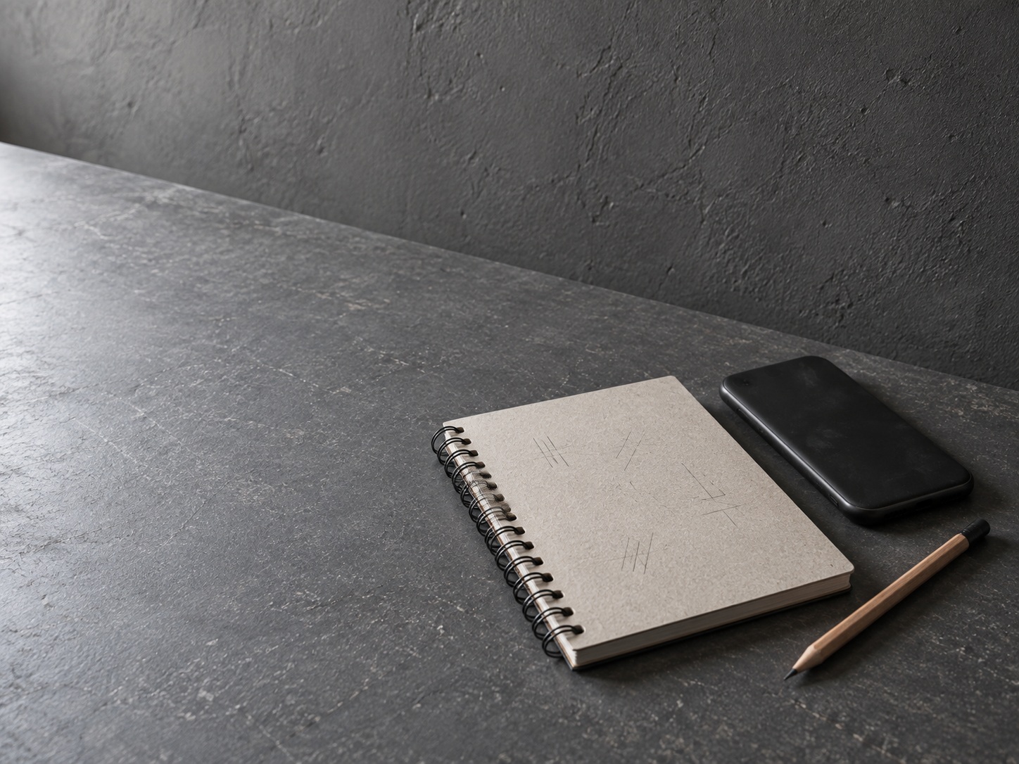 Un cuaderno de espiral cerrado con marcas de lápiz apenas legibles en la portada, un lápiz y un teléfono boca abajo sobre un escritorio gris pizarra.