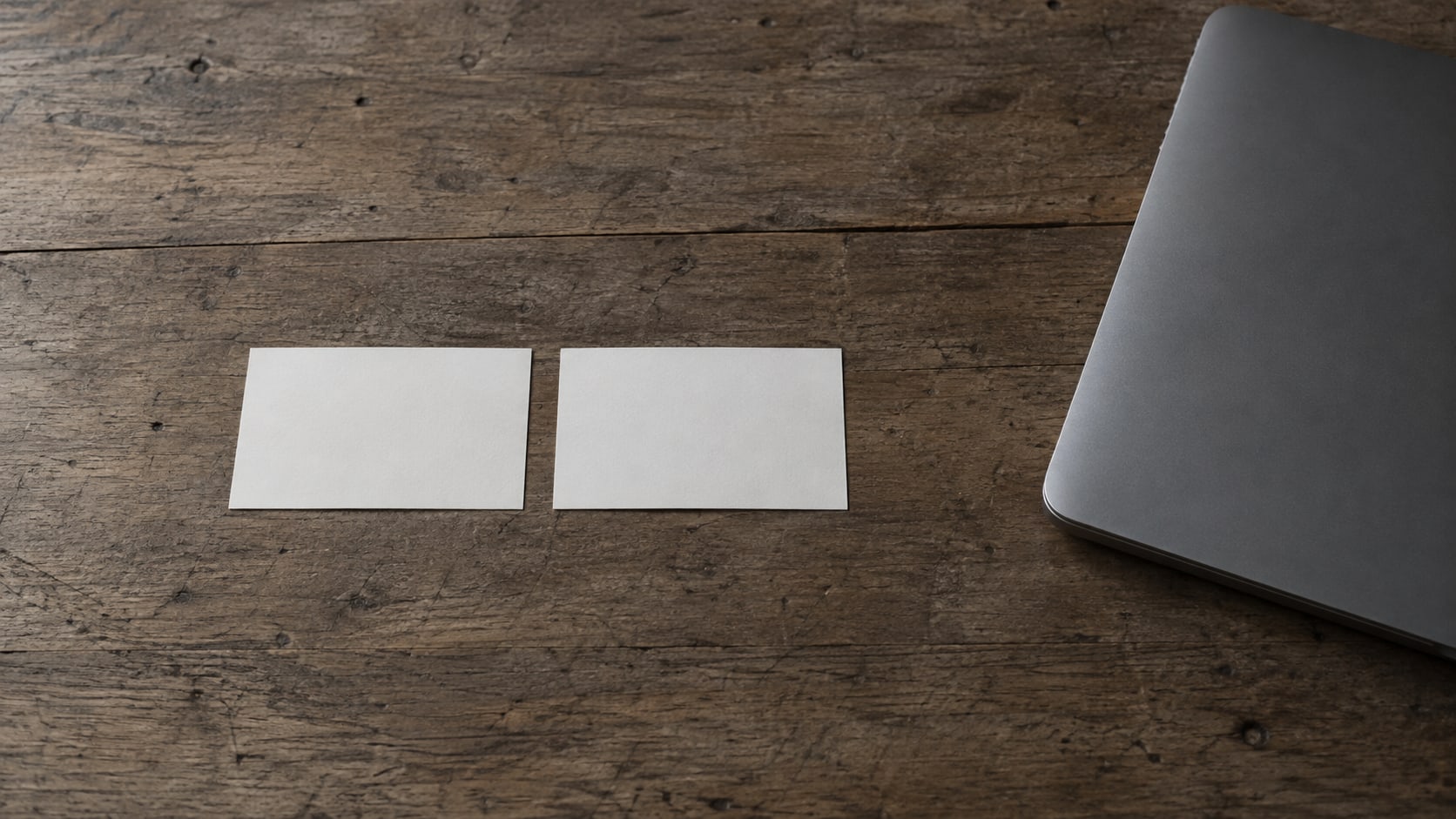Two identical blank index cards side by side on a plain wooden desk beside a closed laptop.