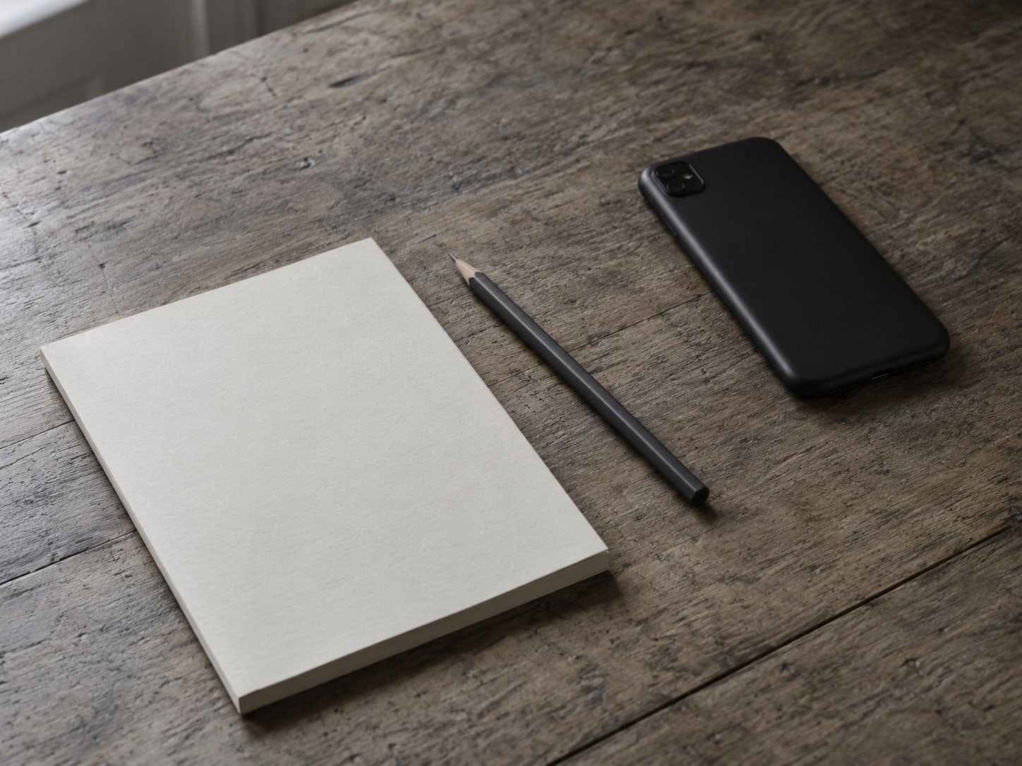 A pencil and a blank notepad beside a switched-off phone lying face down on a desk.