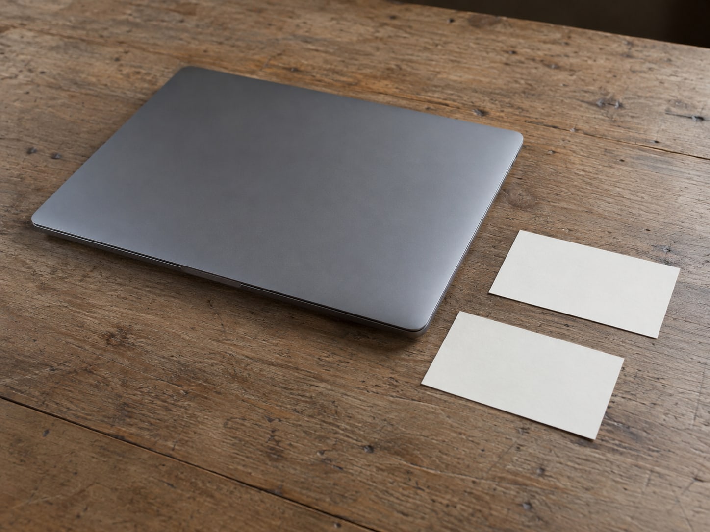A closed laptop beside two identical blank index cards on a plain wooden desk.