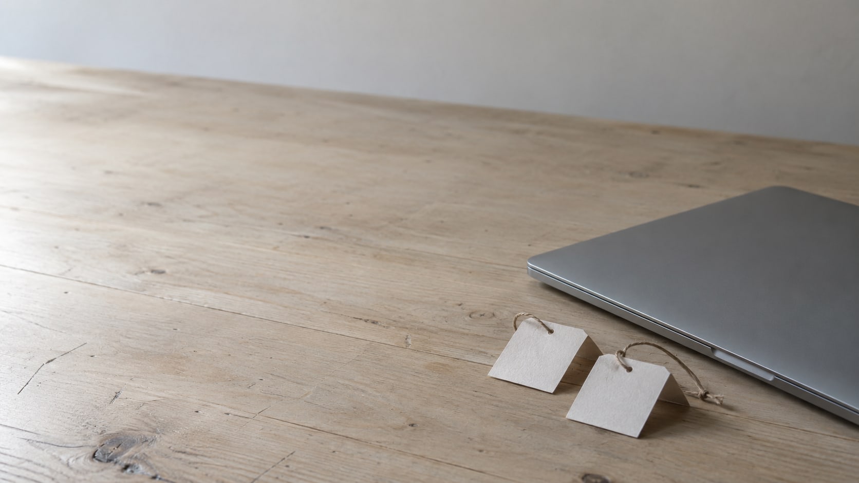A closed laptop beside two folded paper tags tied with string on a plain wooden desk.