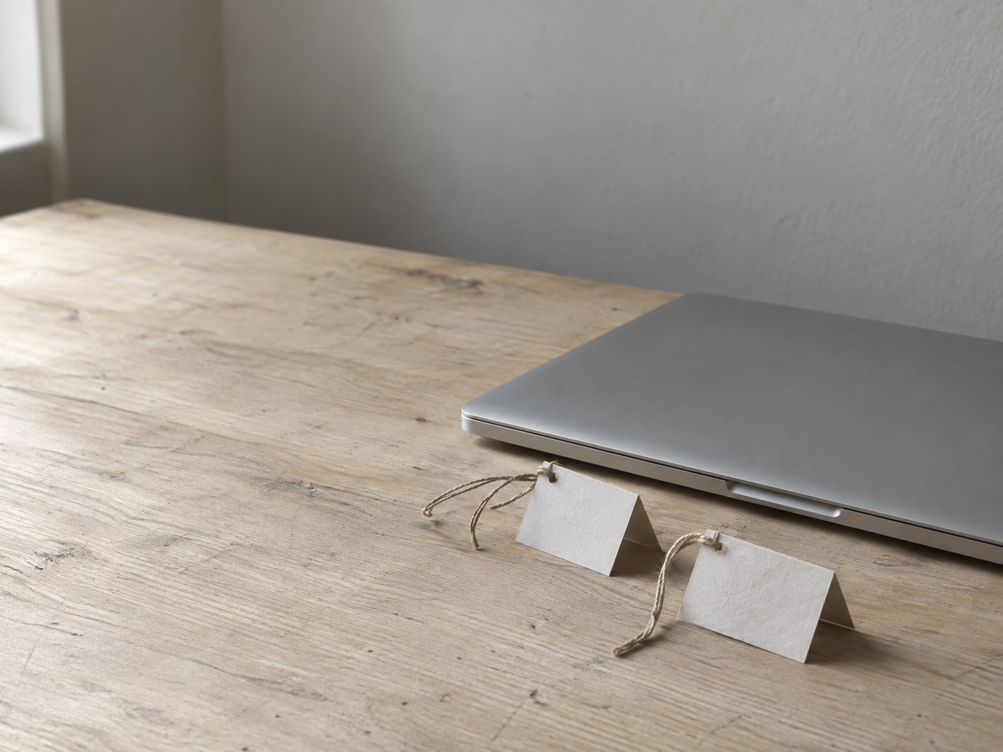 A closed laptop beside two folded paper tags tied with string on a plain wooden desk.