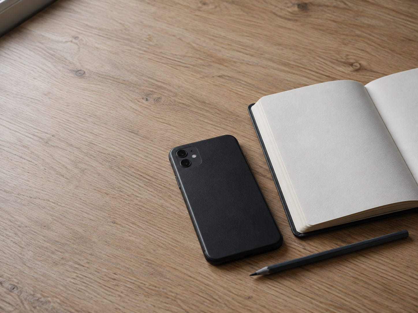 A switched-off phone lying face down beside an open blank notebook and a pencil on a light oak desk.
