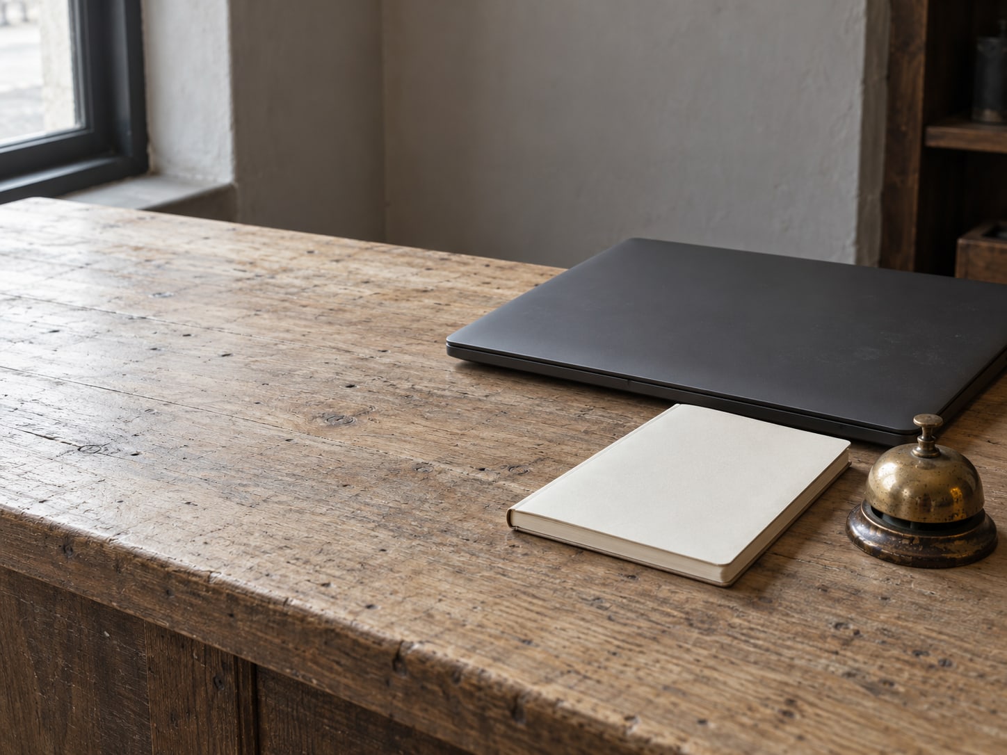 A closed laptop beside a blank appointment book and a small bell on a shop counter.