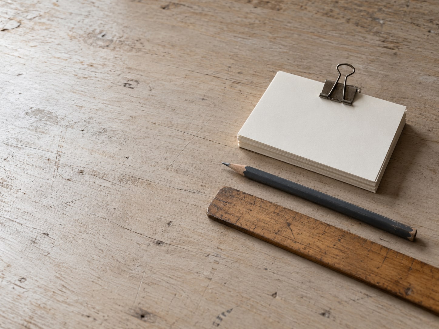 A small stack of index cards held with a binder clip beside a pencil and a worn wooden ruler on a desk.