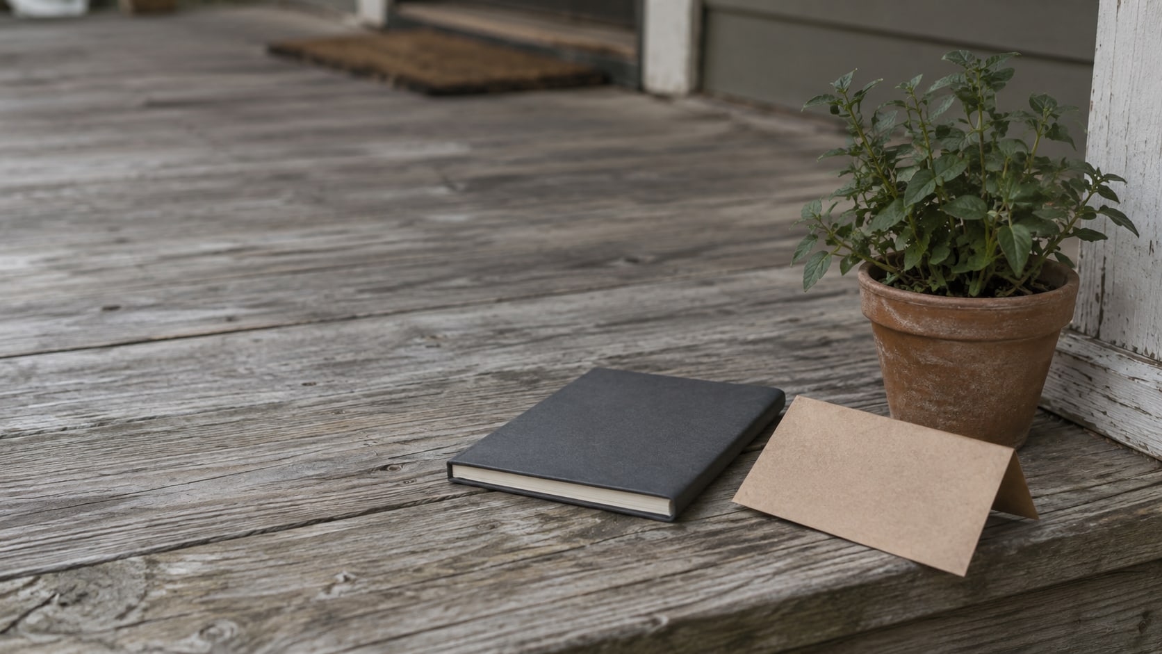 A closed notebook, a folded blank door hanger and a small potted plant on a wooden porch step in soft morning light.