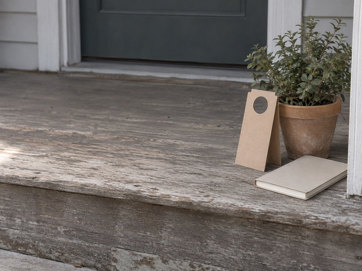 A wooden front porch step with a folded blank door hanger, a small potted plant and a closed notebook in soft morning light.